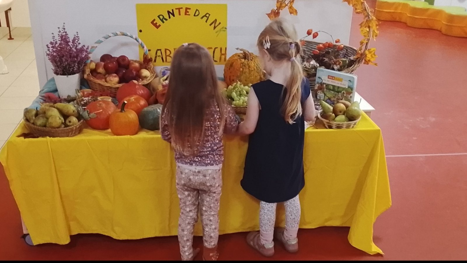 2 Mädchen stehen mit Rücken zur Kamera an einem Tisch mit Äpfeln, Nüssen etc. 