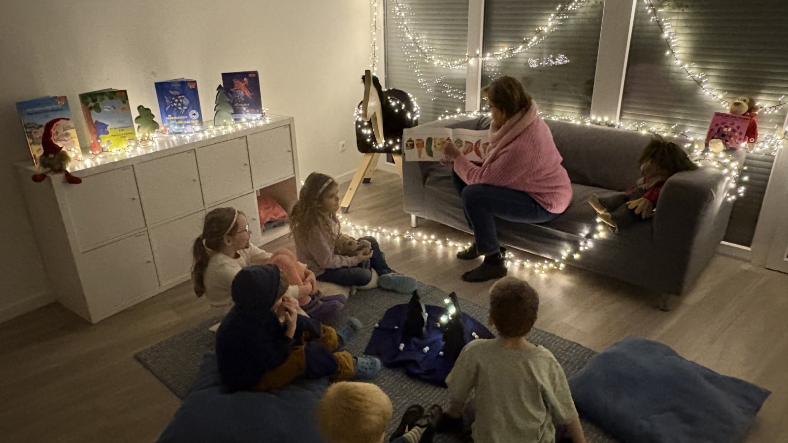 Kindergruppe mit dem Rücken zur Kamera in einem mit Lichterkette beleuchteten Raum lauschend beim Vorlesen